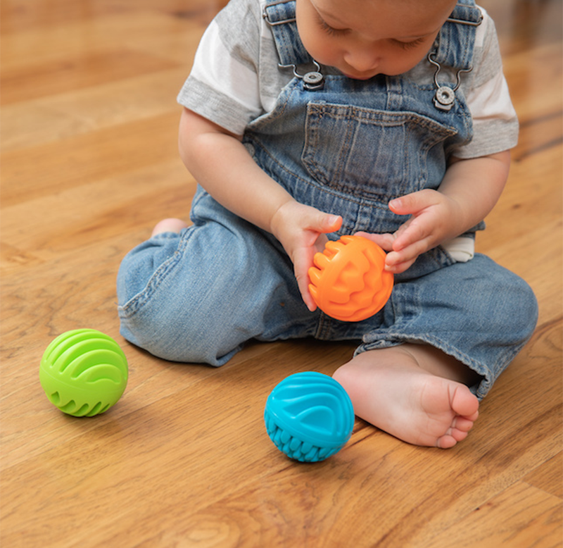 A young child in denim overalls sits on a wooden floor, examining an orange textured ball; green and blue balls nearby create a playful, curious scene.