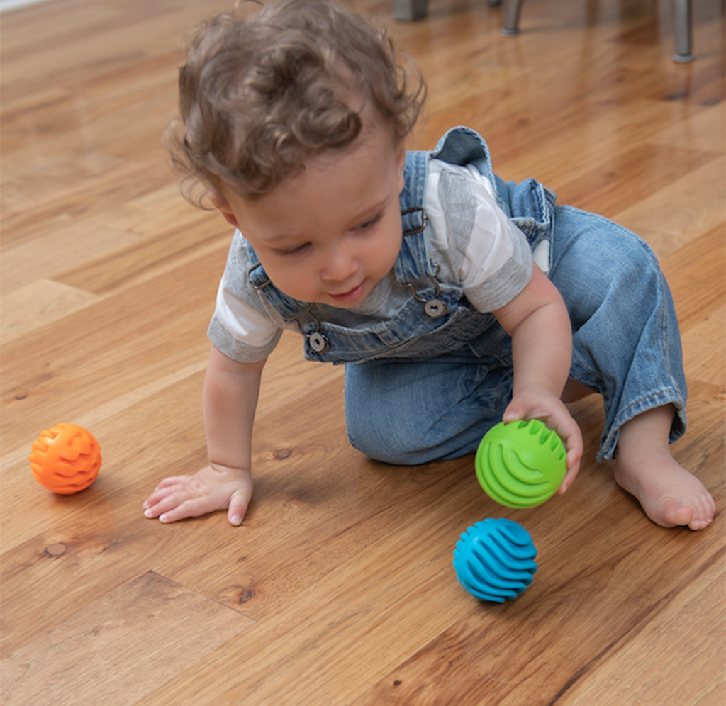 A curly-haired toddler in denim overalls kneels on a wooden floor, reaching for a green textured ball beside blue and orange toys; playful, curious mood.