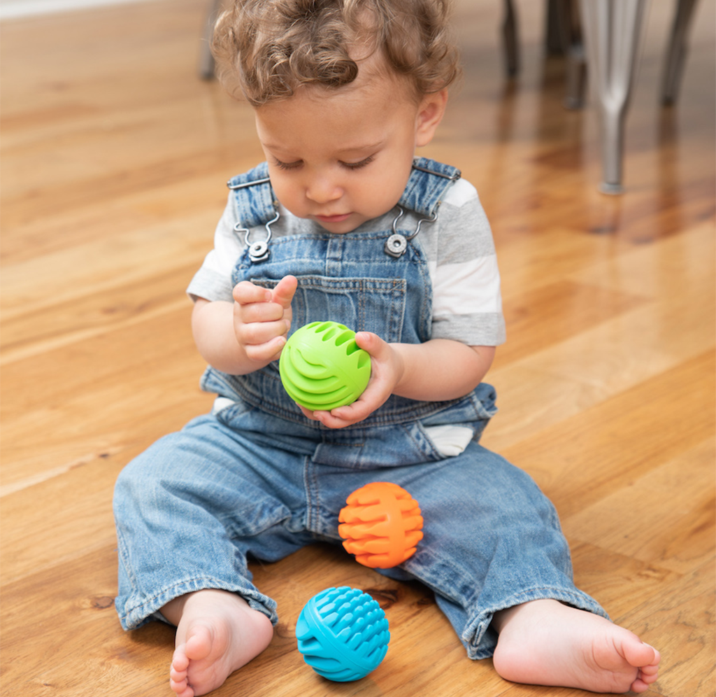 A toddler in denim overalls sits barefoot on a wooden floor, studying a bright green sensory ball, with orange and blue textured balls nearby.
