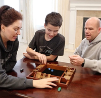 A woman, teenage boy, and bald man lean over a wooden tabletop puzzle with dice, smiling and discussing moves in a bright, relaxed room.