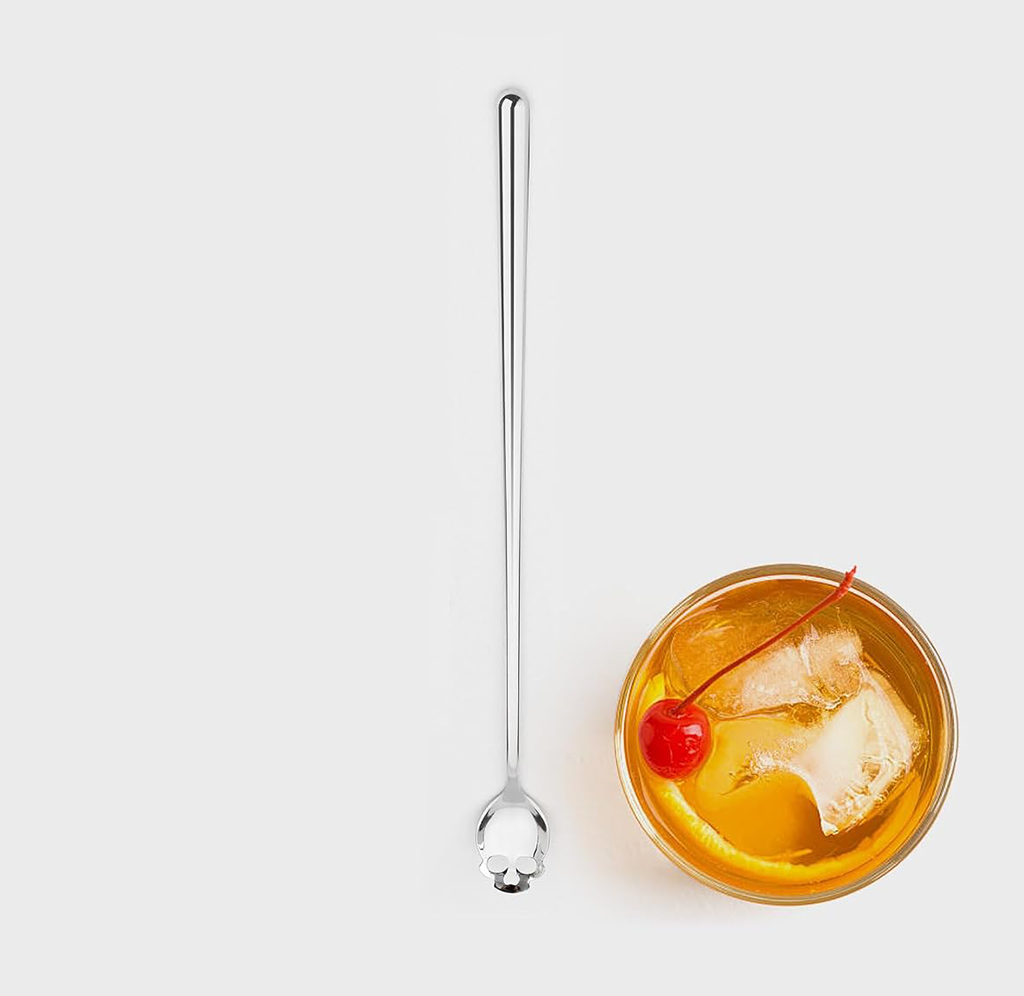 A long, slender stainless steel cocktail spoon with a decorative skull detail at the end, shown next to a glass of orange cocktail with ice, a lemon slice, and a cherry on a light gray background.