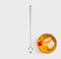 A long, slender stainless steel cocktail spoon with a decorative skull detail at the end, shown next to a glass of orange cocktail with ice, a lemon slice, and a cherry on a light gray background.