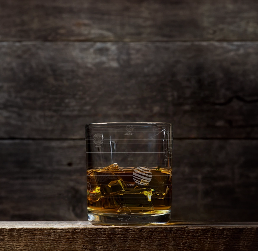 A small glass tank with yellow fish and a striped round fish sits on a dark wooden shelf against a shadowy wall, creating a quiet, moody scene.