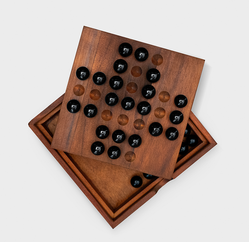 A wooden solitaire peg game with glossy black and translucent brown marbles sits atop its open storage tray, arranged diagonally against a plain white background.