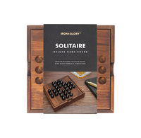 A boxed Iron & Glory wooden Solitaire deluxe game board, shown with a walnut-colored peg board and black glass marbles; refined, premium presentation.