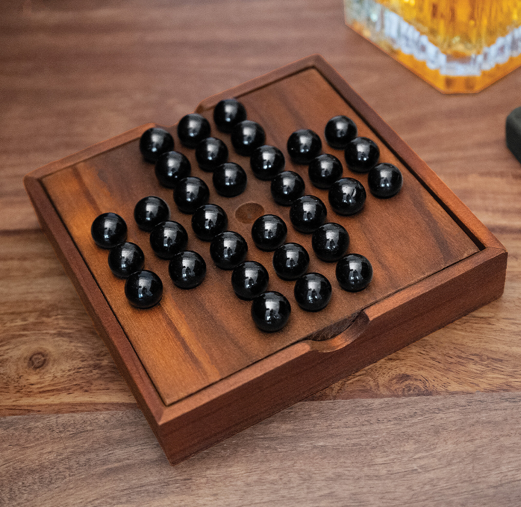 A wooden solitaire board holds glossy black marbles in a cross-like pattern, with a few empty holes; a warm, inviting tabletop scene.