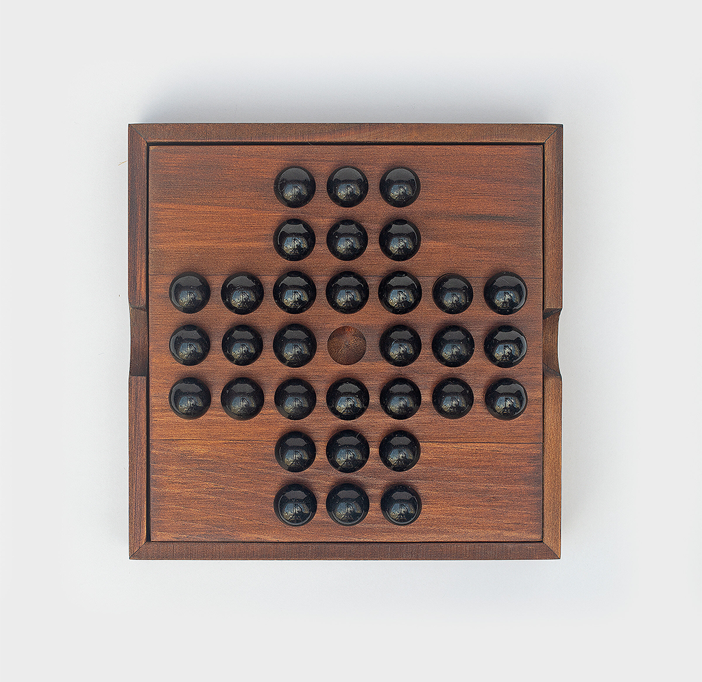 A wooden tabletop peg solitaire board with black marbles in a cross-shaped pattern and one empty center hole, photographed against white; calm, handcrafted feel.