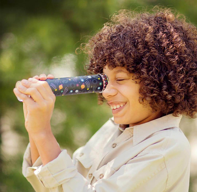 A smiling child with curly hair looks through a colorful kaleidoscope outdoors, enjoying a playful moment against a softly blurred green background.
