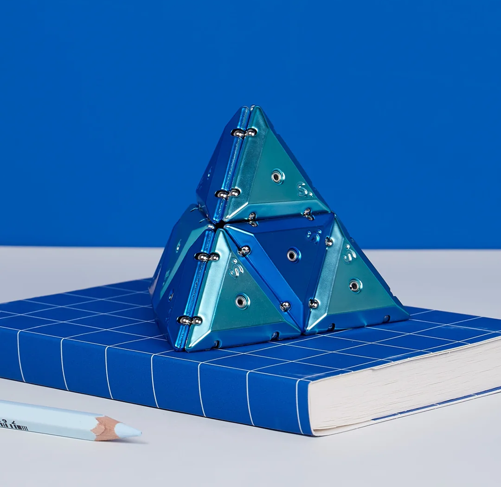 Geode chrome magnetic triangular pieces assembled into a pyramid, sitting on top of a book and next to a pencil. 