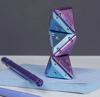 A purple and blue metallic geometric notebook stand made of interlocking triangular panels, shown balancing on top of a lined notebook with a purple pen nearby on a gray and white background.