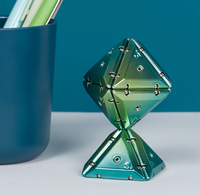 A metallic green and blue iridescent geometric magnetic puzzle styled on a white surface next to a blue cup.