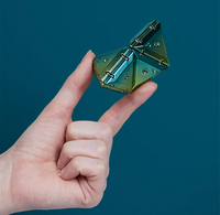 A hand holding a small metallic geometric puzzle toy with a green-to-blue iridescent finish against a solid dark teal background.