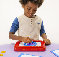 A smiling child uses a marker on a red magnetic drawing board, creating colorful shapes at a table; a playful, focused learning moment.