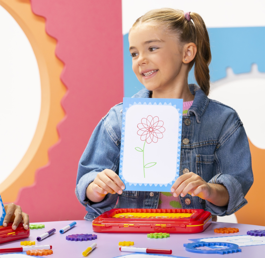A smiling girl in a denim jacket proudly displays a flower drawing at a colorful craft table, with markers and art supplies around her.