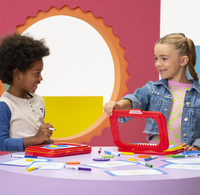 Two smiling children share colorful art supplies at a table; a girl opens a red pencil case as a boy holds a marker in a bright, playful classroom.