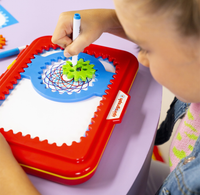 A child uses a blue marker and green gears in a red Spirograph toy to draw colorful looping patterns, showing focused creative play.