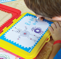 A child uses a blue marker to write “Robot” on a colorful magnetic drawing board, surrounded by robot sketches; focused, playful creativity.