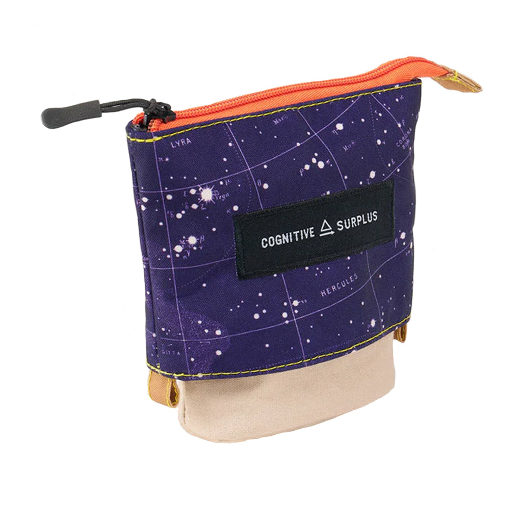A small zippered pouch with a dark purple starry space print, an orange zipper, a black brand patch reading 'COGNITIVE SURPLUS', and a beige fabric flap at the bottom, shown on a white background.