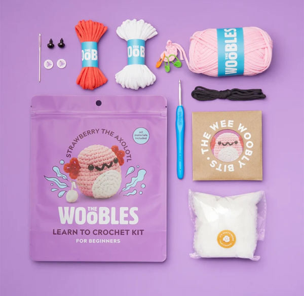 A beginner crochet kit arranged on a purple background, with yarn, hooks, safety eyes, stuffing, and a strawberry axolotl project pouch; colorful, playful packaging.