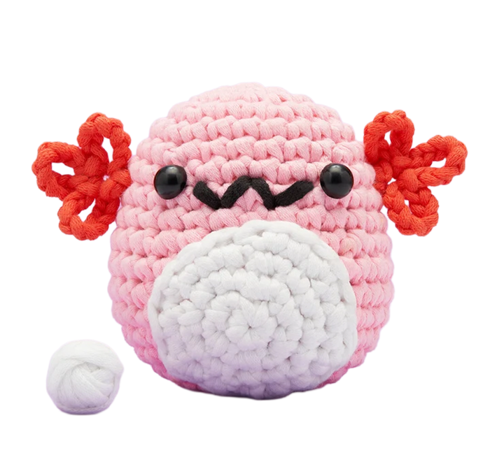 A cheerful pink crocheted axolotl plush with red frilly gills, black eyes, a stitched smile, and a white belly sits beside a yarn ball.