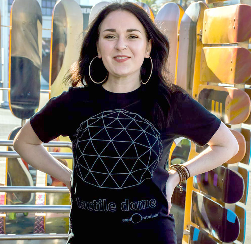 A person is wearing the tactile dome t-shirt. The shirt features a reflective geodesic dome with "Tactile Dome" and "Exploratorium" printed in reflective ink below.
