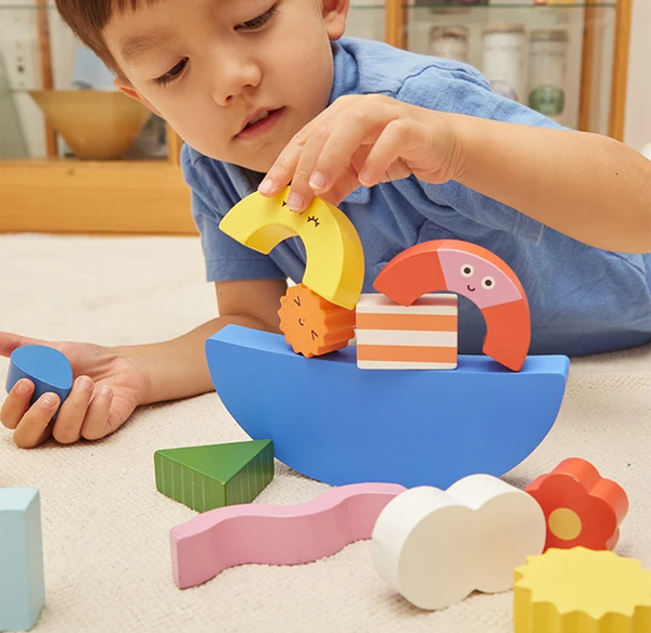 A focused young boy in a blue shirt builds a colorful wooden toy boat, carefully placing a yellow crescent beside smiling shapes; playful, calm scene.
