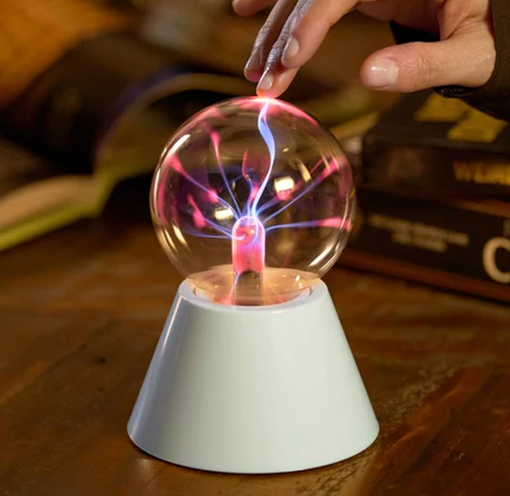 A plasma globe lamp with a white conical base sits on a wooden table, with a hand touching the glass sphere to create colorful electric arcs. Books are blurred in the background.