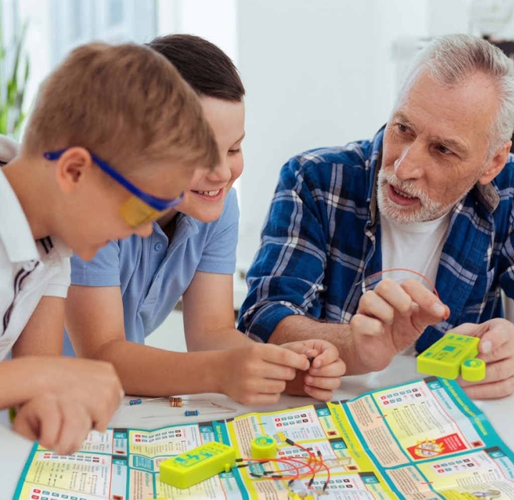 A smiling grandfather and two children build an electronic circuit together at a table, leaning over a colorful instruction sheet in a bright room.