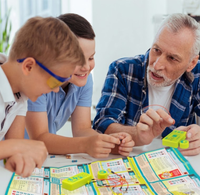 A smiling grandfather and two children build an electronic circuit together at a table, leaning over a colorful instruction sheet in a bright room.