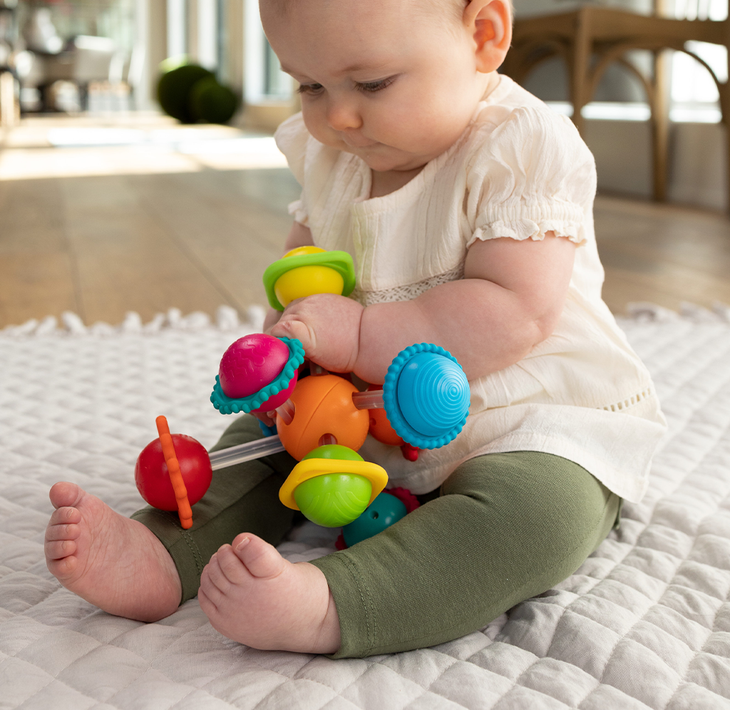 A baby in a white top and green leggings sits on a quilt, intently exploring a colorful spinning toy in a calm, sunlit room.