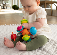A baby in a white top and green leggings sits on a quilt, intently exploring a colorful spinning toy in a calm, sunlit room.