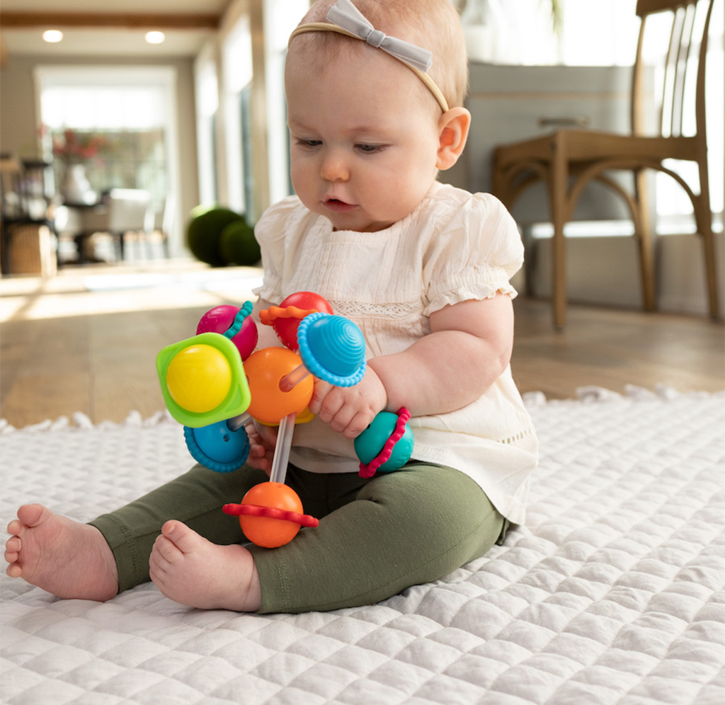 A curious baby in a white dress and gray headband sits on a quilted mat, exploring a colorful toy in a bright, cozy home.
