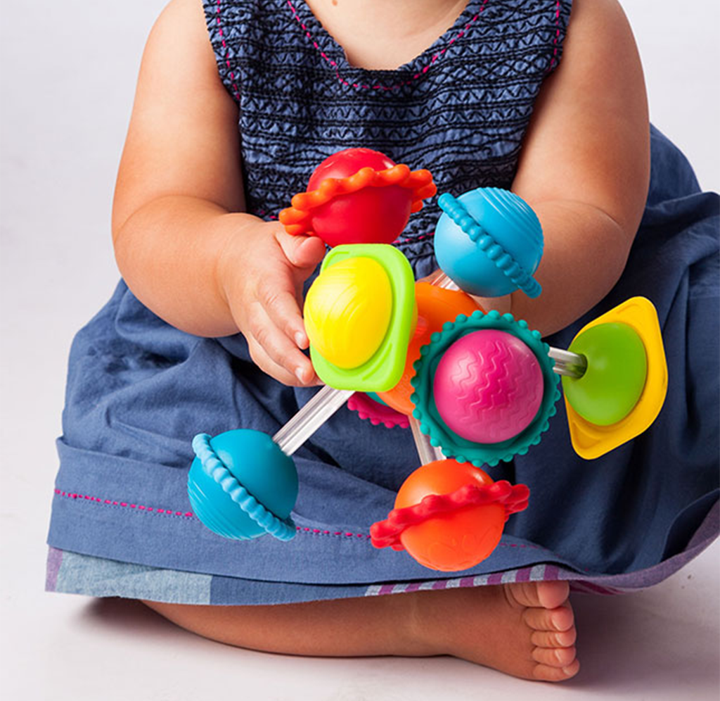 A seated baby in a blue dress holds a bright, colorful spinning toy with textured red, blue, green, and pink parts, conveying playful curiosity.