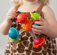 A young child in a giraffe-print apron holds a colorful cluster of suction-cup toys, exploring their shapes with curiosity and playful delight.
