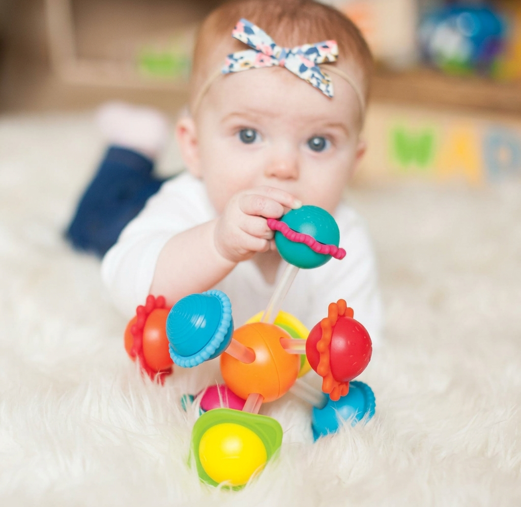 A curious baby wearing a floral bow lies on a soft white rug, chewing a colorful teething toy; the bright scene feels playful and gentle.