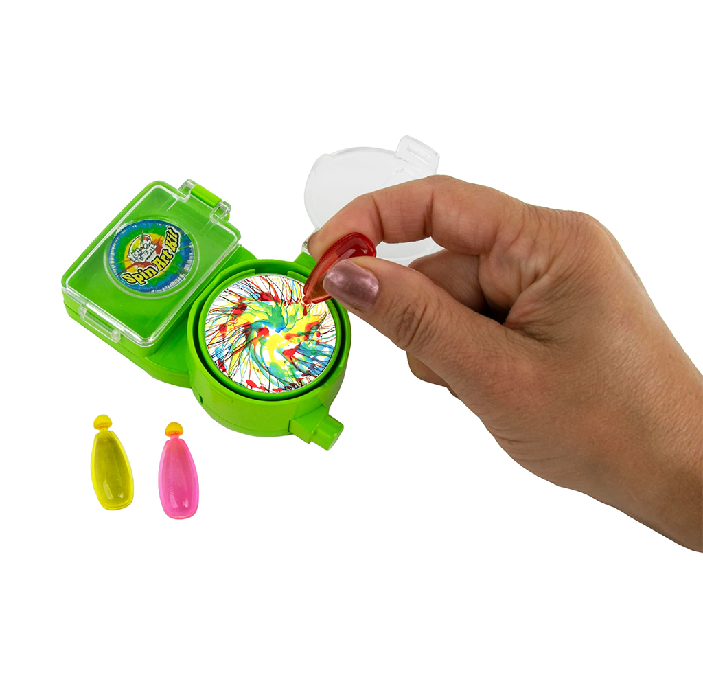 A hand uses a green spin-art toy to drip red paint onto a colorful circular design, with yellow and pink paint bottles nearby, suggesting playful creativity.