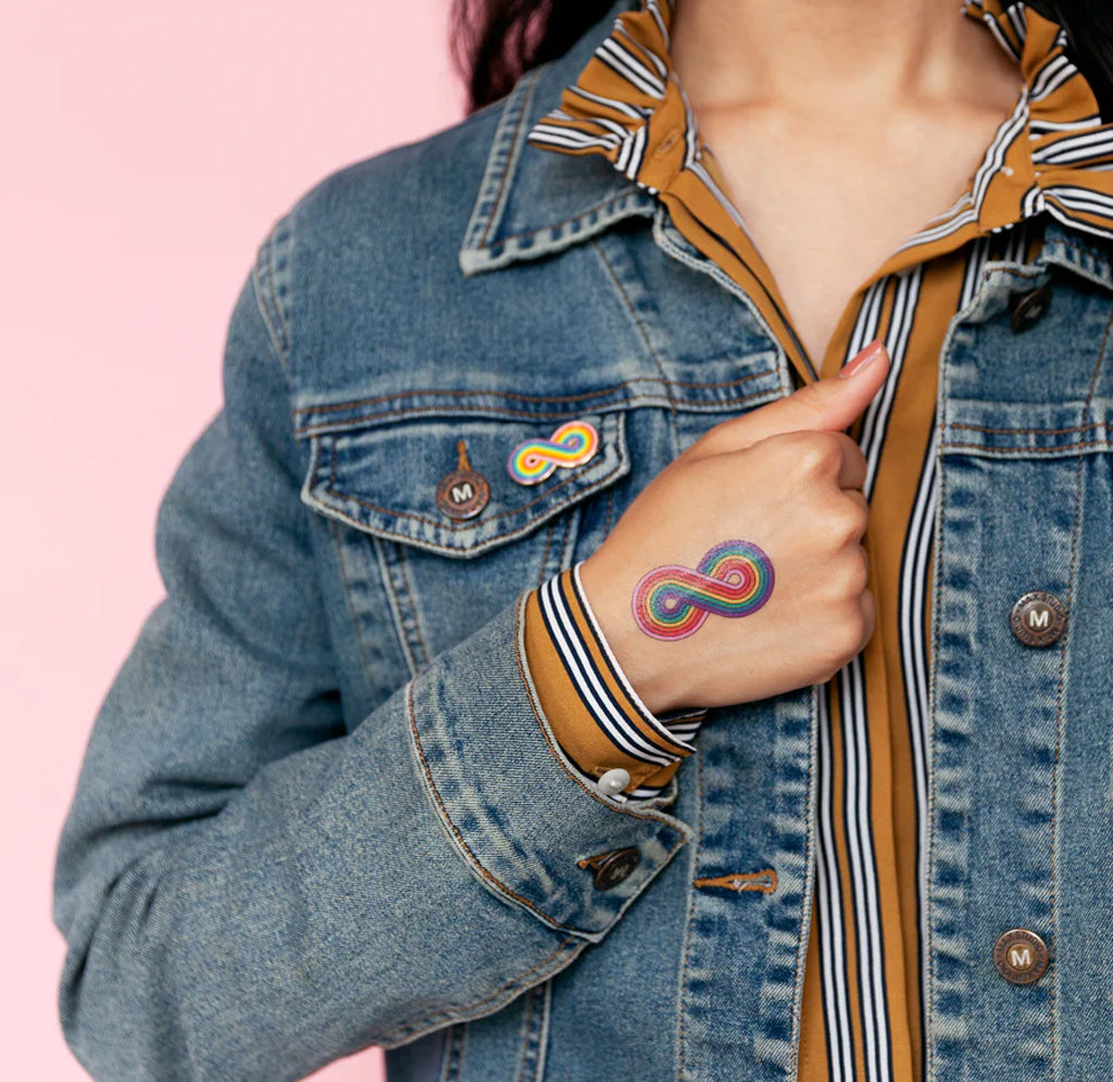 A person is holding their hand in front of their chest, the back of their hand has a rainbow infinity tattoo on it. 