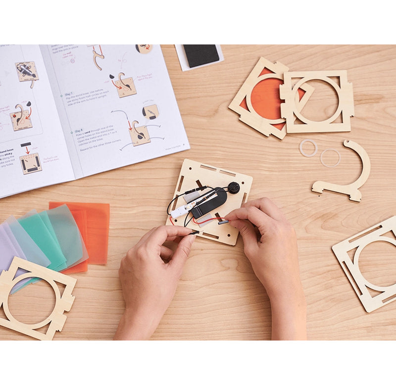 Hands assembling a small electronic device on a wooden table with a manual and additional components.