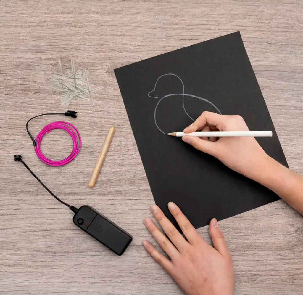 Person drawing a duck on a black sheet of paper with a pen, surrounded by electronic components on a wooden surface.