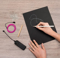 Person drawing a duck on a black sheet of paper with a pen, surrounded by electronic components on a wooden surface.