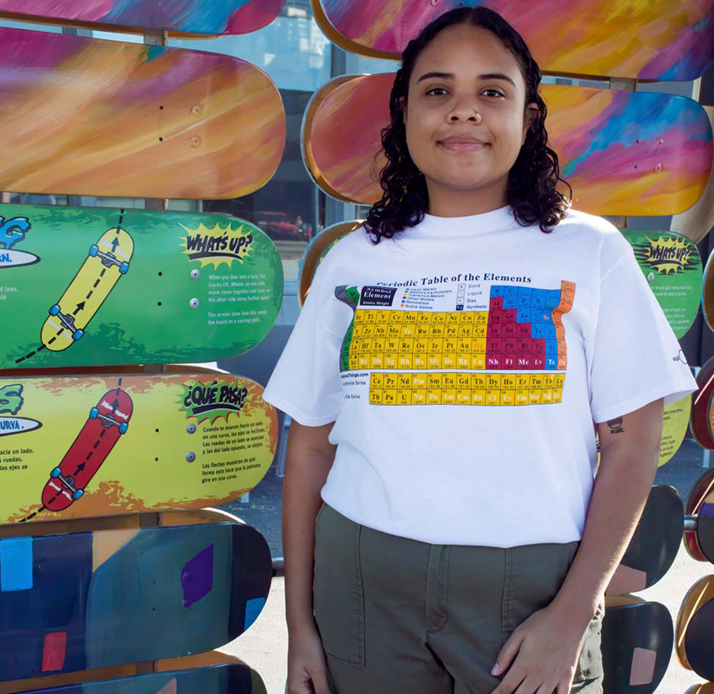 A woman wearing a white shirt with the periodic table of elements of the front. She is standing in front of a colorful skateboard exhibit at the Exploratorium.
