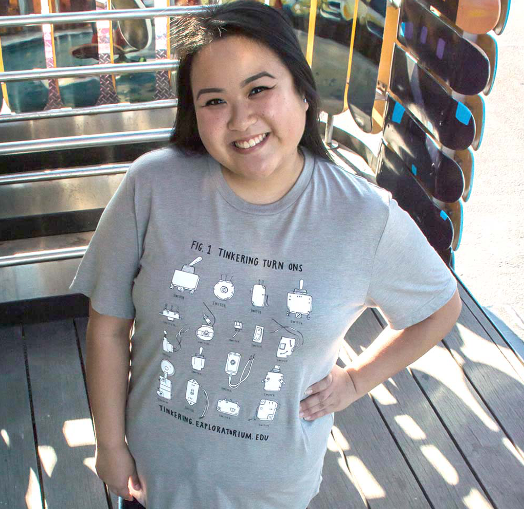 A young woman is standing in a skateboarding exhibit; the thinking t-shirt is gray with different switches that turn things on in white.
