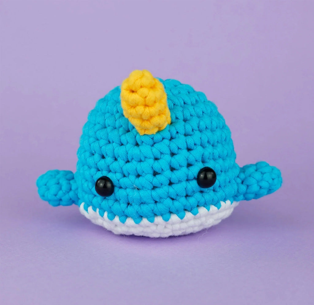 A crochet blue and white narwhal on a purple background. 