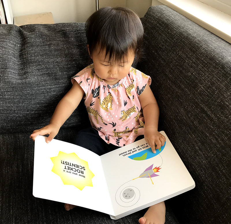 A toddler sits on a couch interacting with the "Rocket Science for babies" book. On the page is a rocketship above Earth and circling the moon. 
