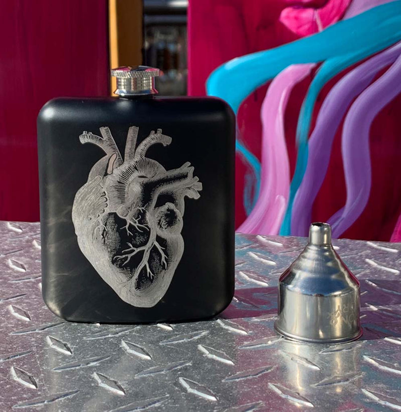 Black coated stainless steel hip flask with a silver anatomical heart on the front. A silver funnel sits to the left. The mouthpiece at the top of the flask is stainless steel. The flask sits on top of a skateboard exhibit.