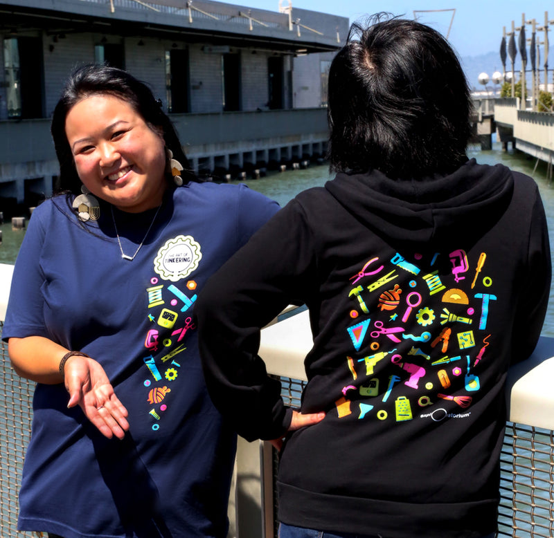 Two individuals are standing on a bridge, one facing forward and one with her back toward the viewer, the woman facing is wearing a blue t-shirt with colorful tools cascading down the front of the shirt, and the other woman is wearing a black hoodie with colorful tools for making on the back.