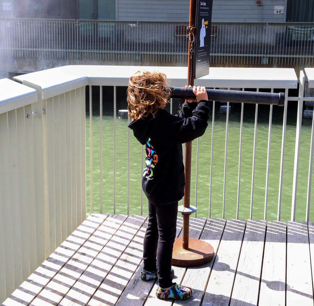 A young child is standing on a pier holding a telescope in her hand, looking to the left. She is wearing the black Art of Tinkering hoodie with brightly colored neon tools in pink, orange, teal, green and yellow.