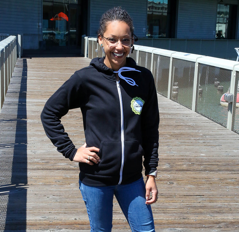 A person is standing in the middle of a bridge and wearing the black Art of Tinkering hoodie with a round gear logo that is white, yellow, and black.A woman is standing on a bridge and wearing a black hoodie with the Art of Tinking logo shaped like a gear in yellow, black, and white.