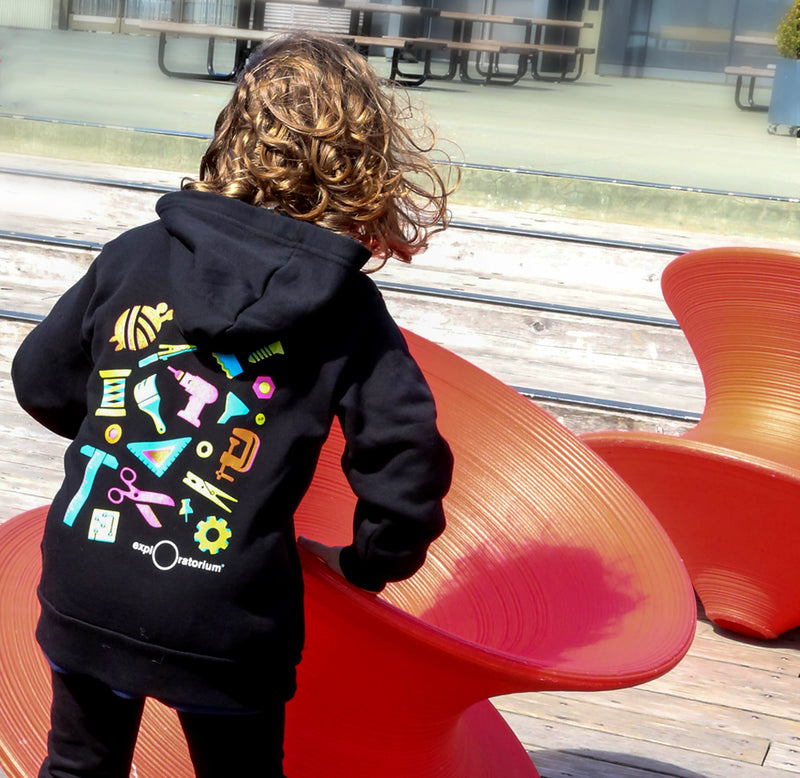 A young child is standing on a pier with her back facing forward. She is wearing the black Art of Tinkering hoodie with brightly colored neon tools in pink, orange, teal, green and yellow.