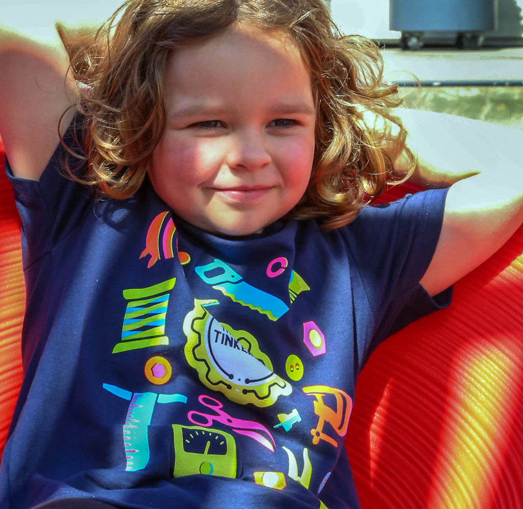 A young child is sitting on a red chair; she is wearing a blue shirt with neon tools in green, teal, pink, orange, and yellow on the front surrounding the gear-shaped Art of Tinkering logo in yellow, white, and black. 